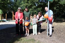 Vier Erwachsene und zwei Kinder durchschneiden gemeinsam ein grünes Eröffnungsband vor einem mit Luftballons geschmückten Baum. Im Hintergrund sind die Spielgeräte des neuen Spielplatzes „Baumschätze“ zu sehen. Rechts steht Oberbürgermeister Torsten Ruban-Zeh, links Frau Steiniger vom Planungsbüro 360° Landschaftsarchitekten.