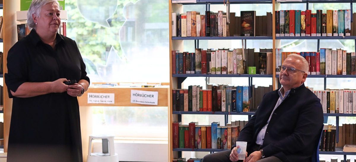 Eine Frau steht in einer Bibliothek zwischen Bücherregalen und spricht, ein Mann sitzt ihr auf einem Stuhl zugewandt und hört zu.