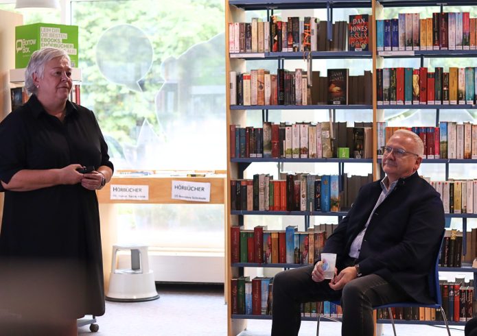 Eine Frau steht in einer Bibliothek zwischen Bücherregalen und spricht, ein Mann sitzt ihr auf einem Stuhl zugewandt und hört zu.