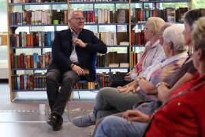 Ein Mann sitzt vor einem Bücherregal auf einem Stuhl und spricht zu mehreren Zuhörerinnen und Zuhörern, die ihm im Halbkreis gegenübersitzen.