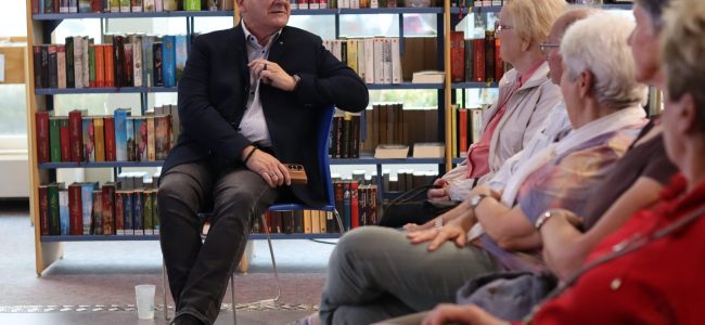Ein Mann sitzt vor einem Bücherregal auf einem Stuhl und spricht zu mehreren Zuhörerinnen und Zuhörern, die ihm im Halbkreis gegenübersitzen.