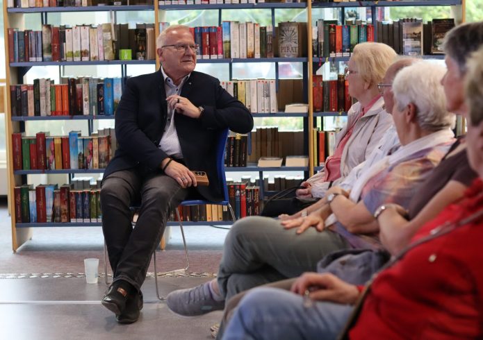 Ein Mann sitzt vor einem Bücherregal auf einem Stuhl und spricht zu mehreren Zuhörerinnen und Zuhörern, die ihm im Halbkreis gegenübersitzen.