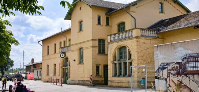 Zweigeschossiges Bahnhofsgebäude im Gründerzeitstil mit ockerfarbenem Putz, grünen Fensterrahmen und grüner Eingangstür. An der Fassade hängt eine runde Bahnhofsuhr. Am rechten Gebäudeflügel befinden sich Bauzäune, dahinter ist ein Wandbild auf einer Bauwand zu erkennen. Im Vordergrund links sitzen zwei Personen auf einer Holzbank, links oben ragt ein Baum ins Bild.