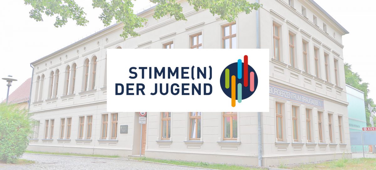 Blick auf das Bürgerzentrum Hoyerswerda, Braugasse 1, ein helles mehrstöckiges Gebäude mit einem zweisprachigen Schriftzug auf Deutsch und Sorbisch. Im Vordergrund hängt ein Straßenschild mit der Aufschrift „Stimme(n) der Jugend" und dem dazugehörigen farbigen Logo auf dunklem Kreishintergrund.