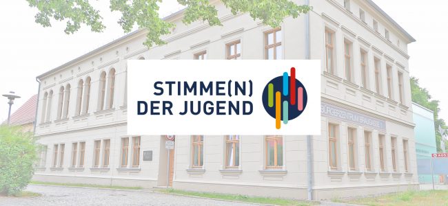 Blick auf das Bürgerzentrum Hoyerswerda, Braugasse 1, ein helles mehrstöckiges Gebäude mit einem zweisprachigen Schriftzug auf Deutsch und Sorbisch. Im Vordergrund hängt ein Straßenschild mit der Aufschrift „Stimme(n) der Jugend" und dem dazugehörigen farbigen Logo auf dunklem Kreishintergrund.