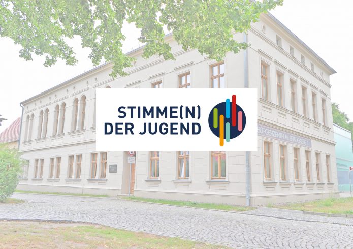 Blick auf das Bürgerzentrum Hoyerswerda, Braugasse 1, ein helles mehrstöckiges Gebäude mit einem zweisprachigen Schriftzug auf Deutsch und Sorbisch. Im Vordergrund hängt ein Straßenschild mit der Aufschrift „Stimme(n) der Jugend" und dem dazugehörigen farbigen Logo auf dunklem Kreishintergrund.