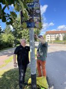 Torsten Ruban-Zeh und Christian Völker-Kieschnick stehen an einem Laternenmast und entfernen einen Aufkleber. Über ihnen hängt ein Plakat zur Aktion „Hoywoj putzt sich“.