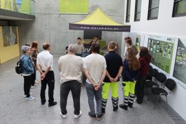 Eine Gruppe junger Menschen und Erwachsener steht in einem Innenbereich vor einem grünen Pavillon und hört einem Sprecher zu. An der Wand sind Informationstafeln zu Strukturwandelprojekten in Hoyerswerda zu sehen.
