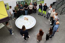 Aus der Vogelperspektive ist eine Gruppe zu sehen, die im Kreis um einen großen runden Tisch steht. Auf dem Tisch befinden sich eine Stadtumrissgrafik, Karten mit Bezeichnungen verschiedener Verwaltungsbereiche, Wasserflaschen und Gebäck.