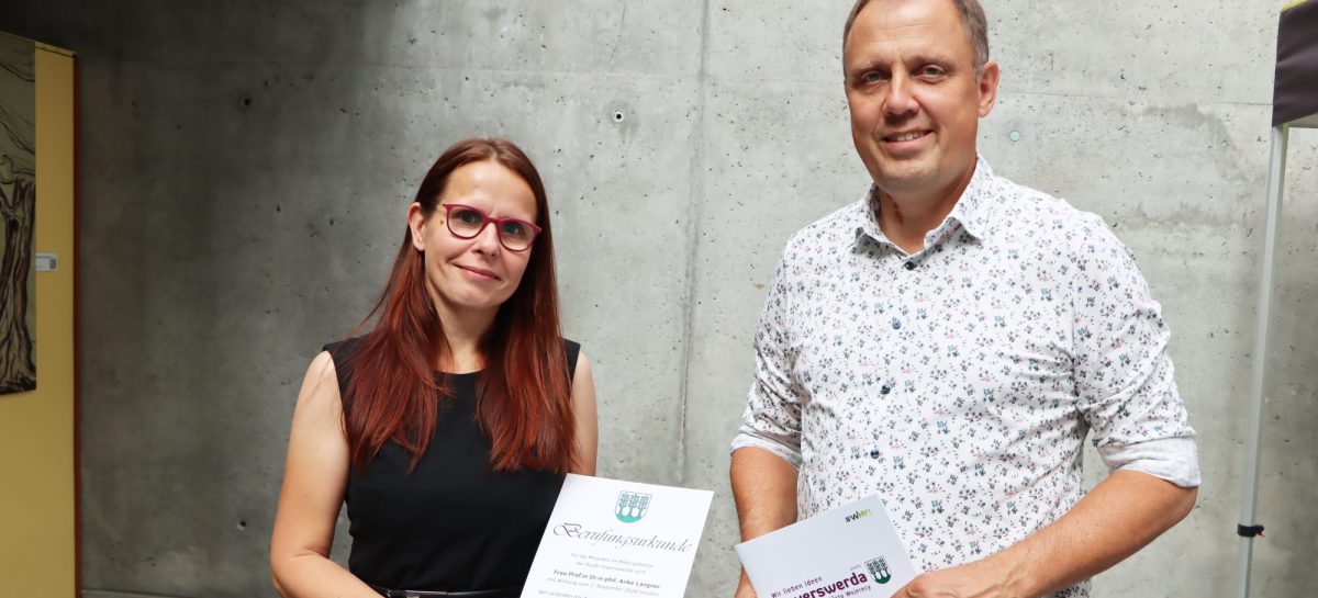 Prof. Dr. Anke Langner und Bürgermeister Mirko Pink stehen vor einer Betonwand. Anke Langner hält die Berufungsurkunde in den Händen, Mirko Pink eine Mappe der Stadt Hoyerswerda.