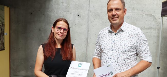 Prof. Dr. Anke Langner und Bürgermeister Mirko Pink stehen vor einer Betonwand. Anke Langner hält die Berufungsurkunde in den Händen, Mirko Pink eine Mappe der Stadt Hoyerswerda.