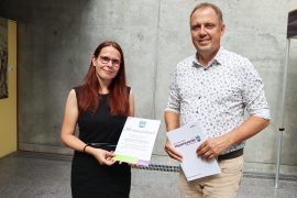 Prof. Dr. Anke Langner und Bürgermeister Mirko Pink stehen vor einer Betonwand. Anke Langner hält die Berufungsurkunde in den Händen, Mirko Pink eine Mappe der Stadt Hoyerswerda.