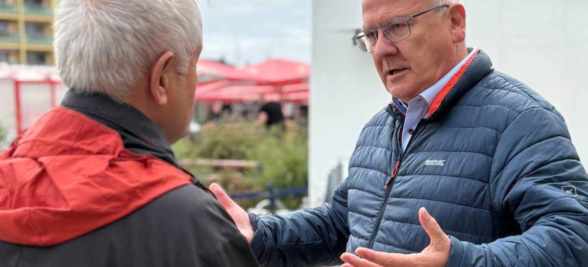 Zwei Männer stehen sich im Freien gegenüber und unterhalten sich. Der rechte Mann trägt eine dunkelblaue Steppjacke und Brille und gestikuliert erklärend mit beiden Händen, der linke Mann im Vordergrund ist von hinten zu sehen und trägt eine rot-schwarze Jacke. Im Hintergrund sind Hochhausfassaden, rote Sonnenschirme und Bäume zu erkennen.
