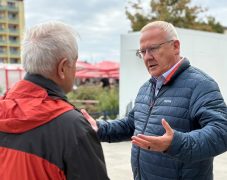 Zwei Männer stehen sich im Freien gegenüber und unterhalten sich. Der rechte Mann trägt eine dunkelblaue Steppjacke und Brille und gestikuliert erklärend mit beiden Händen, der linke Mann im Vordergrund ist von hinten zu sehen und trägt eine rot-schwarze Jacke. Im Hintergrund sind Hochhausfassaden, rote Sonnenschirme und Bäume zu erkennen.