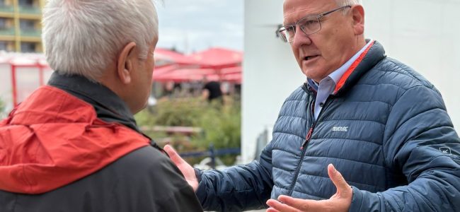 Zwei Männer stehen sich im Freien gegenüber und unterhalten sich. Der rechte Mann trägt eine dunkelblaue Steppjacke und Brille und gestikuliert erklärend mit beiden Händen, der linke Mann im Vordergrund ist von hinten zu sehen und trägt eine rot-schwarze Jacke. Im Hintergrund sind Hochhausfassaden, rote Sonnenschirme und Bäume zu erkennen.