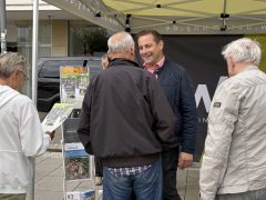 Christian Hoffmann steht lächelnd unter einem Pavillon mit #WHY!-Beschriftung im Gespräch mit mehreren älteren Personen. Eine der Personen hält einen Newsletter mit der Überschrift „Spielplatz eröffnet