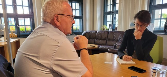Oberbürgermeister Torsten Ruban-Zeh sitzt an einem Tisch und spricht mit Lydia DePillis, die ihm gegenübersitzt und Notizen in einen Schreibblock einträgt.
