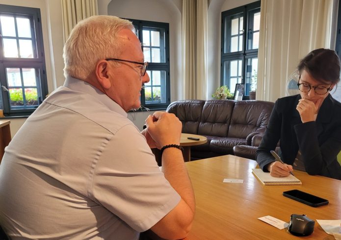 Oberbürgermeister Torsten Ruban-Zeh sitzt an einem Tisch und spricht mit Lydia DePillis, die ihm gegenübersitzt und Notizen in einen Schreibblock einträgt.