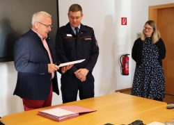 Oberbürgermeister Torsten Ruban-Zeh hält eine Urkunde in den Händen und steht neben einem Feuerwehrbeamten in Uniform. Eine Frau verfolgt die Übergabe.