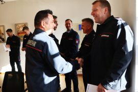 Zwei uniformierte Feuerwehrbeamte schütteln sich lächelnd die Hand. Im Hintergrund stehen weitere Kollegen, teilweise mit Urkunden in den Händen.