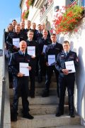 Eine Gruppe uniformierter Feuerwehrbeamter und eine Frau stehen auf einer Außentreppe. Mehrere Beamte halten ihre Urkunden vor sich.
