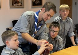 Mehrere Pfadfinder stehen und sitzen an einem Tisch. Ein Betreuer unterstützt einen Jungen bei der Demonstration einer bestimmten Knotentechnik, während weitere Kinder zuschauen.