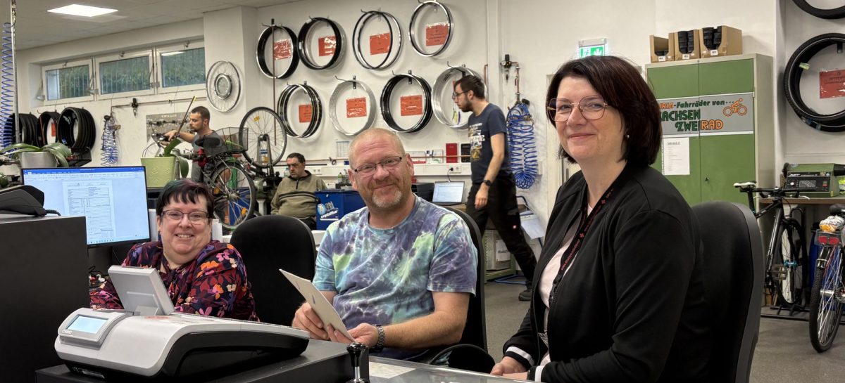 Beate Gröger sitzt gemeinsam mit Bianka Lyssy und Thomas Kloß am Arbeitsplatz im ServiceCenter der Lausitzer Werkstätten. Im Hintergrund sind Fahrräder und Fahrradteile zu sehen.