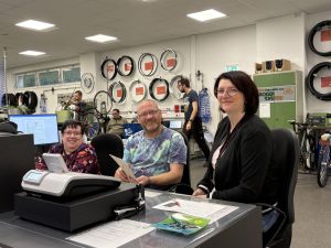 Beate Gröger sitzt gemeinsam mit Bianka Lyssy und Thomas Kloß am Arbeitsplatz im ServiceCenter der Lausitzer Werkstätten. Im Hintergrund sind Fahrräder und Fahrradteile zu sehen.