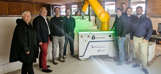 Kathrin Schlesinger, Geschäftsführerin Lautech GmbH, Oberbürgermeister Torsten Ruban-Zeh und eine Gruppe von Projektbeteiligten stehen in einem Baustellenraum rund um den gelben WallBot. Hinter dem Baurobotiksystem ist bereits errichtetes Mauerwerk zu sehen.