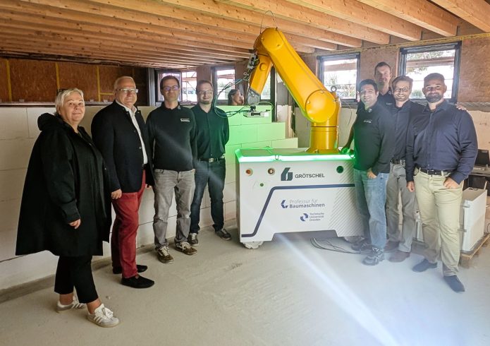 Kathrin Schlesinger, Geschäftsführerin Lautech GmbH, Oberbürgermeister Torsten Ruban-Zeh und eine Gruppe von Projektbeteiligten stehen in einem Baustellenraum rund um den gelben WallBot. Hinter dem Baurobotiksystem ist bereits errichtetes Mauerwerk zu sehen.