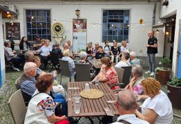 Eine Gruppe von Besucherinnen und Besuchern sitzt und steht in einem begrünten Innenhof an mehreren Tischen. Eine Person informiert die Gruppe über das Unternehmen. Im Hintergrund sind historische Ausstellungsstücke zu sehen.