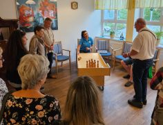 Eine Gruppe von Besucherinnen und Besuchern steht in einem Aufenthaltsraum um einen länglichen Holztisch. Eine Mitarbeiterin in blauer Arbeitskleidung demonstriert ein Spiel mit einer Kugel und kleinen Holzkegeln auf dem Tisch.