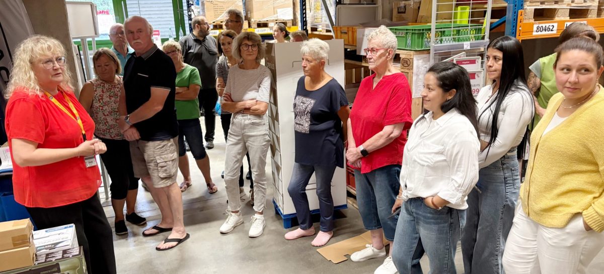 Eine Mitarbeiterin in rotem Shirt spricht zu einer Gruppe von Besucherinnen und Besuchern in einem Warenlager. Um die Gruppe herum stehen hohe Regale mit Kartons und verpackten Waren auf Paletten.
