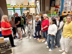 Eine Mitarbeiterin in rotem Shirt spricht zu einer Gruppe von Besucherinnen und Besuchern in einem Warenlager. Um die Gruppe herum stehen hohe Regale mit Kartons und verpackten Waren auf Paletten.