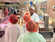 Drei Teilnehmende mit Schutzkleidung und roten Haarnetzen stehen hinter einer Fleischtheke. Ein Mitarbeiter in weißer Arbeitskleidung erklärt ihnen die Abläufe im Arbeitsbereich der Globus Fachmetzgerei.