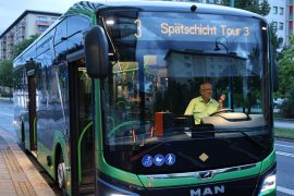 Ein grün-schwarzer Elektrobus steht an einer Haltestelle. Auf der digitalen Anzeige an der Front ist „Spätschicht Tour 3“ zu lesen. Hinter der Frontscheibe sitzt der Busfahrer.