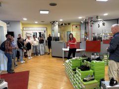 Eine Mitarbeiterin in rotem Shirt erklärt einer Gruppe von Besucherinnen und Besuchern die Arbeit in einem Fachgeschäft für Gesundheitsprodukte. Im Raum sind zahlreiche Schuhkartons, Schuhe sowie Produkte aus den Bereichen Bandagen und Orthesen zu sehen.