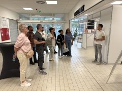 Eine Gruppe von Besucherinnen und Besuchern steht in einem hellen Eingangsbereich und hört einem Mitarbeiter zu. Hinter ihm befinden sich Ausstellungswände und Informationsmaterialien von Unternehmen.