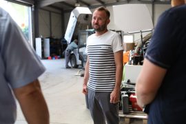 Tommy Schimang steht in einer Werkstatthalle und spricht mit zwei Personen, die am linken und rechten Bildrand angeschnitten zu sehen sind. Im Hintergrund arbeitet ein Mitarbeiter an einer großen Maschine.