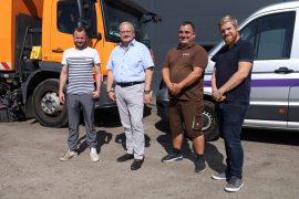 Vier Männer stehen auf dem Betriebsgelände zwischen einem orangefarbenen Straßenreinigungsfahrzeug und einem weißen Transporter. Zu sehen sind Geschäftsführer Tommy Schimang, Oberbürgermeister Torsten Ruban-Zeh, ein Mitarbeiter des Unternehmens und Wirtschaftsförderer Sebastian Schindler.