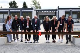 Neun Vertreterinnen und Vertreter der DB Bahnbau Gruppe, des Landkreises und der Kommune stehen hinter einer Schiene. In der Mitte der Schiene ist eine große rote Schleife befestigt.