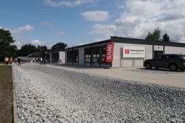 Außenansicht eines lang gestreckten, eingeschossigen Gebäudes. An der Fassade steht „DB Bahnbau Gruppe GmbH – Qualifizierungszentrum Knappenrode“. Mehrere Personen gehen am Gebäude entlang.