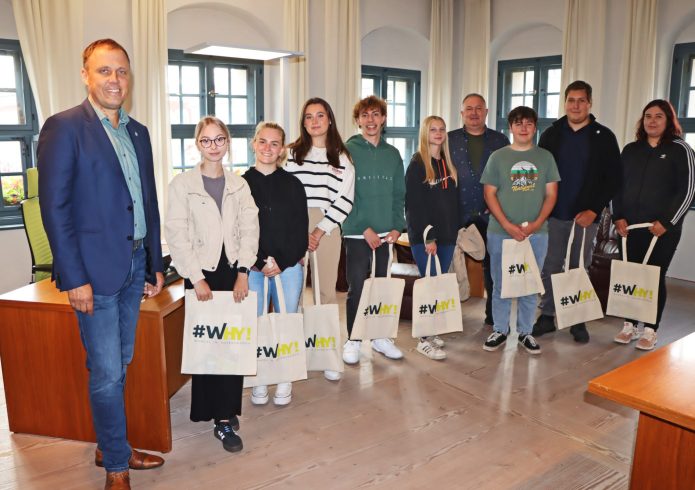 Ein Mann im Anzug steht in einem Amtszimmer neben neun jungen Menschen, die jeweils eine Stofftasche mit der Aufschrift „#WHY! Wandel in Hoyerswerda" in der Hand halten.