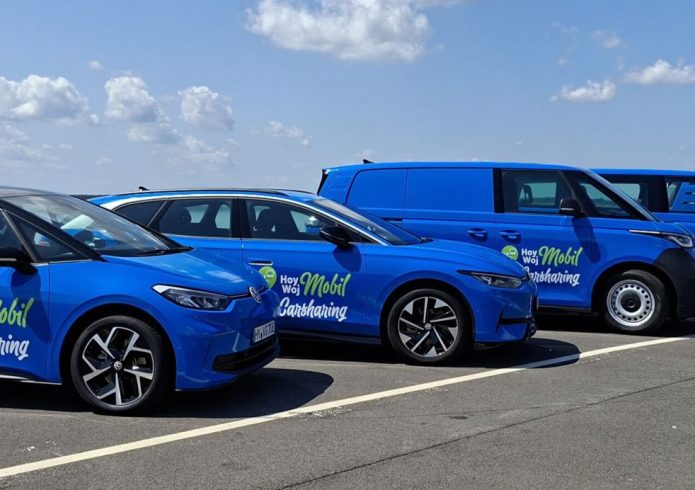 Fünf blaue Hoy Woy Mobil Carsharing-Fahrzeuge stehen nebeneinander auf einem Parkplatz am See.