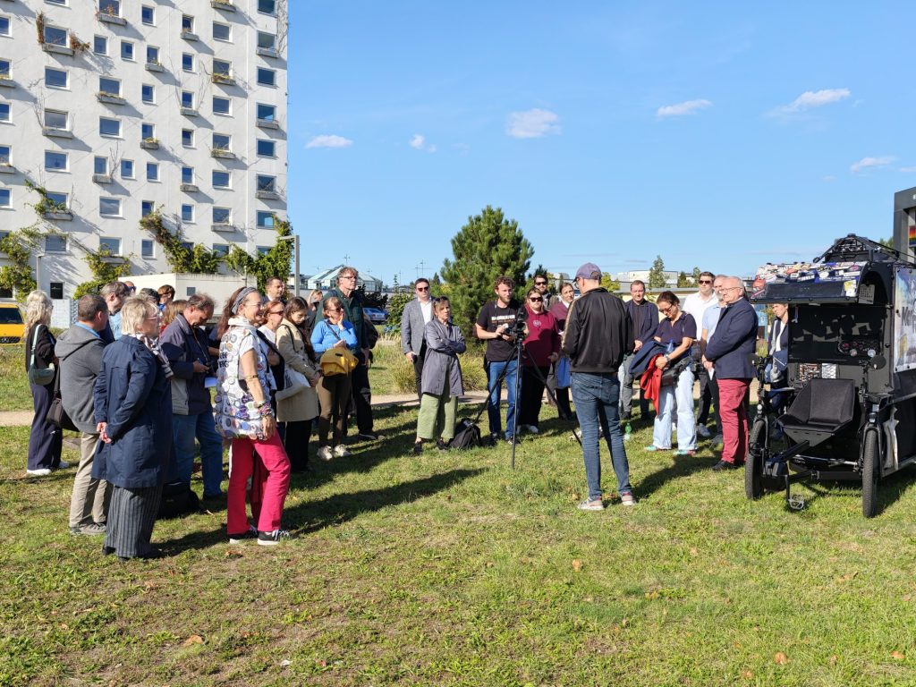 Alternativtext: Eine Gruppe von Menschen steht auf einer Wiese im Zentralpark und hört einem Mann zu, der mit dem Rücken zur Kamera steht. Rechts sind der Oberbürgermeister und ein Lastenrad mit Veranstaltungstechnik zu sehen.