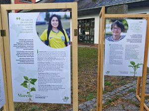 Zwei Ausstellungstafeln in Holzrahmen zeigen Porträts von Frauen und Texte über ihr Leben in Hoyerswerda. Links ist eine Frau im gelben Fußballtrikot zu sehen, rechts eine Frau mit Brille.