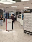 Eine Frau spricht an einem Rednerpult mit Mikrofon im Lausitz-Center. Neben ihr steht eine Tafel mit dem Titel „See my Story. Copyright by Madgermanes“, dahinter sind weitere Ausstellungstafeln zu sehen.