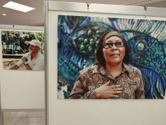 Zwei fotografische Porträts auf Ausstellungstafeln: Im Vordergrund ist eine Frau mit Brille und einer Hand auf der Brust vor einem farbigen Wandbild zu sehen. Links dahinter hängt das Porträt eines Mannes mit heller Kappe.