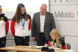 Eine Sportakrobatin sitzt am Tisch und unterschreibt im aufgeschlagenen Goldenen Buch. Neben ihr stehen ihre Partnerin mit einer Goldmedaille und Oberbürgermeister Torsten Ruban-Zeh und schauen zu.