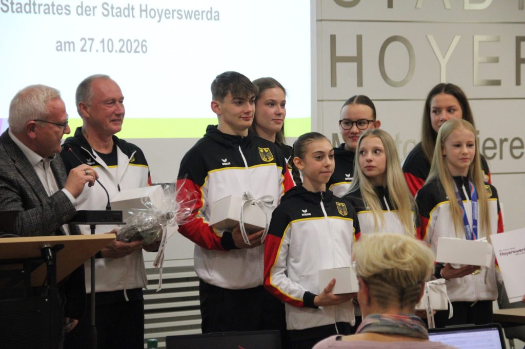 Oberbürgermeister Torsten Ruban-Zeh spricht am Rednerpult. Neben ihm stehen Trainer Sergej Jeriomkin und sieben Sportakrobatinnen und Sportakrobaten in schwarz-rot-weißen Trainingsanzügen. Mehrere halten Geschenkverpackungen, zwei tragen Medaillen.